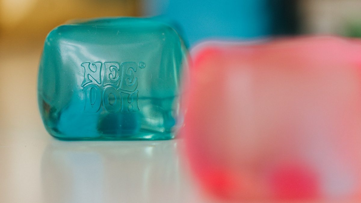 Turkis NeeDoh Nice Cube med preget logo på et bord, en rosa kube uskarp i bakgrunnen
